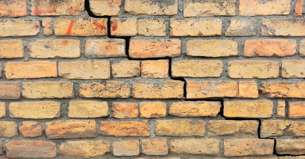 Close-up of a wall of tan and orange bricks. A stair-step crack follows a diagonal mortar line across several rows.