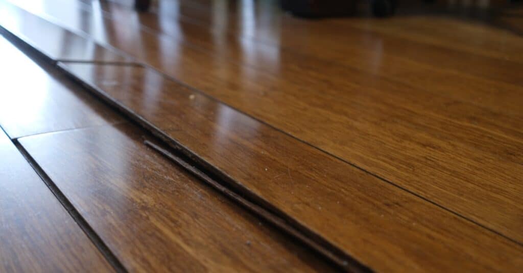 Close-up of an uneven wood floor that reflects light. One row of boards is raised, and the next row is coming loose.