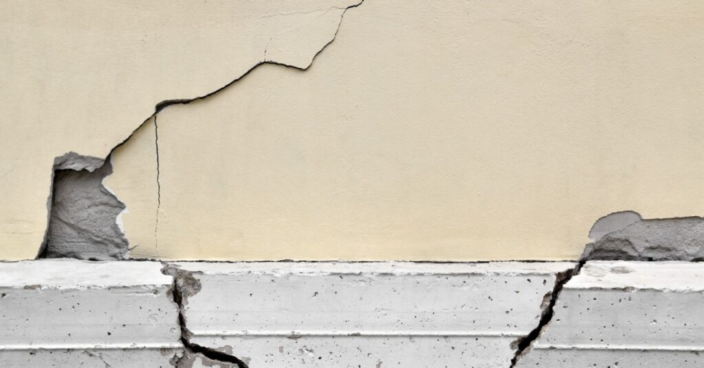 An exterior wall is above a concrete foundation with cracks and broken edges. A long, jagged crack runs across the wall.