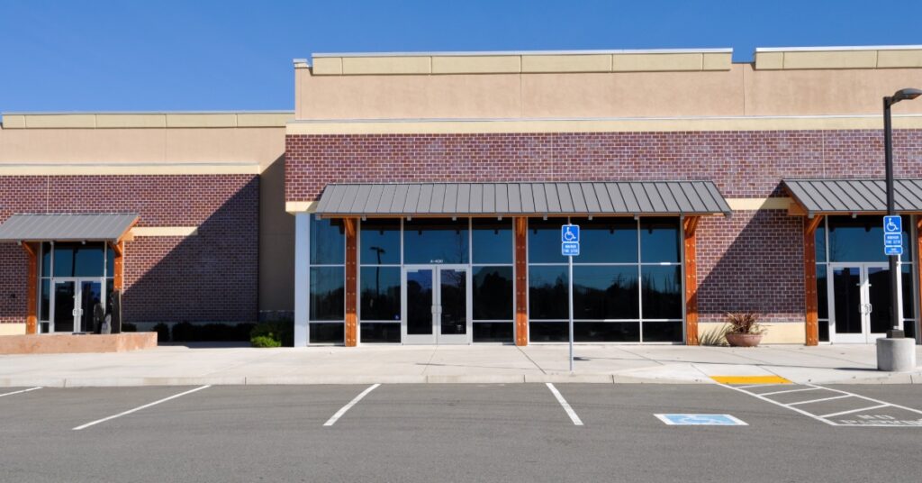 A parking lot is in front of a commercial storefront with brick walls, large glass windows, and metal awnings.