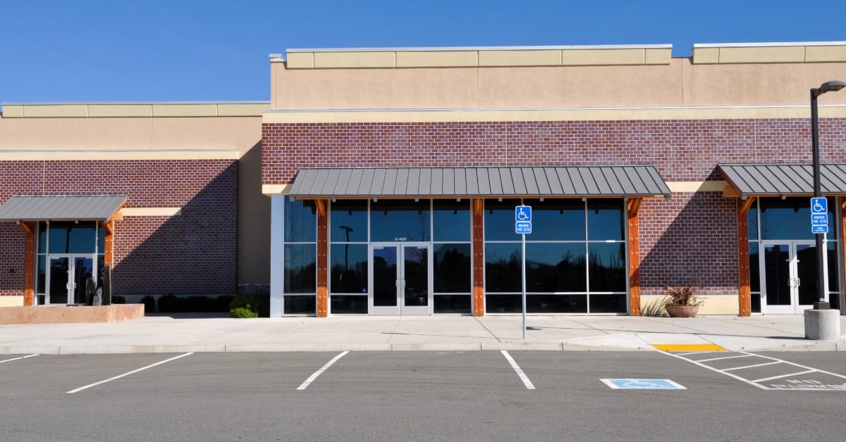 A parking lot is in front of a commercial storefront with brick walls, large glass windows, and metal awnings.