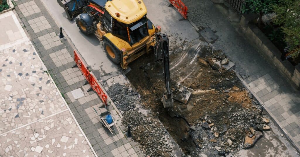 A yellow excavator works on a street excavation between sidewalks and parked cars. The road surface is broken open.