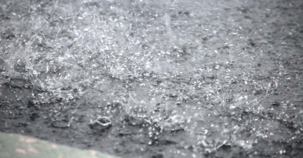 Heavy rain splashes on a dark paved surface, creating bright droplets across the ground. A curb is in the foreground.