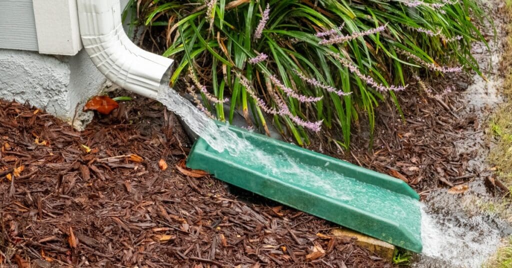A downspout releases water onto a green splash block by a house. Mulch and purple flowering plants surround the block.