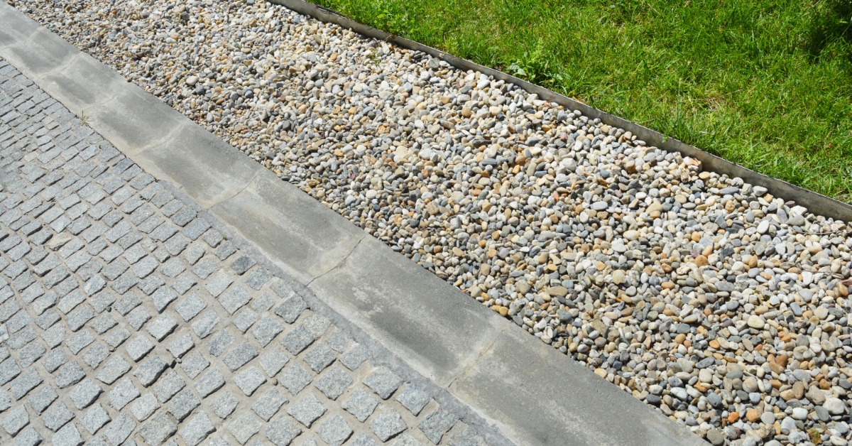 A gravel drainage bed sits between a paved path and a grassy yard. Edging keeps the gravel out of the grass.