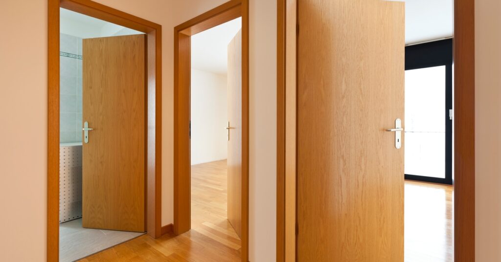 A hallway has three open wooden doors with matching wood trim. The floor is wood, and one door leads to a tiled room.