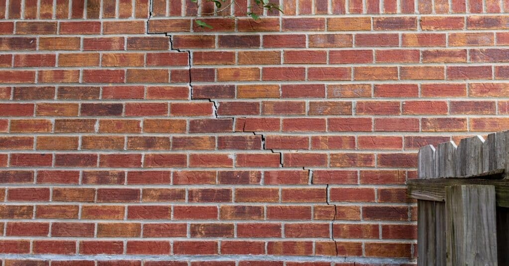 A red brick wall has a stair-step crack through several mortar lines. A gray wooden fence is perpendicular to the wall.