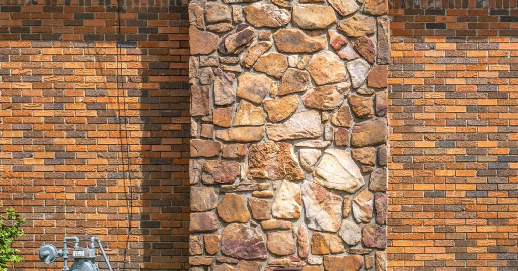 Straight-on view of a stone chimney against a brick exterior wall. Utility meters and a small wall vent are next to it.