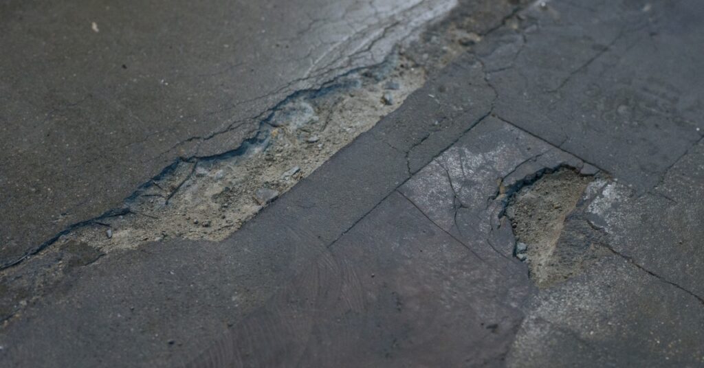 Cracks of various sizes run diagonally across dark concrete. Spalling forms rough, shallow cavities beside the crack.