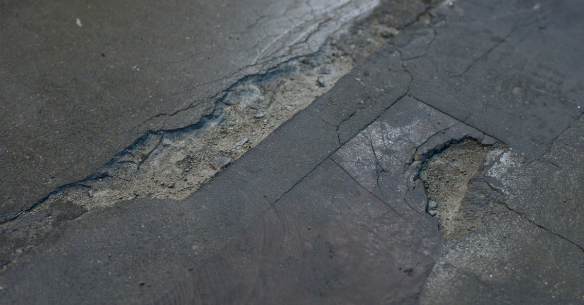 Cracks of various sizes run diagonally across dark concrete. Spalling forms rough, shallow cavities beside the crack.
