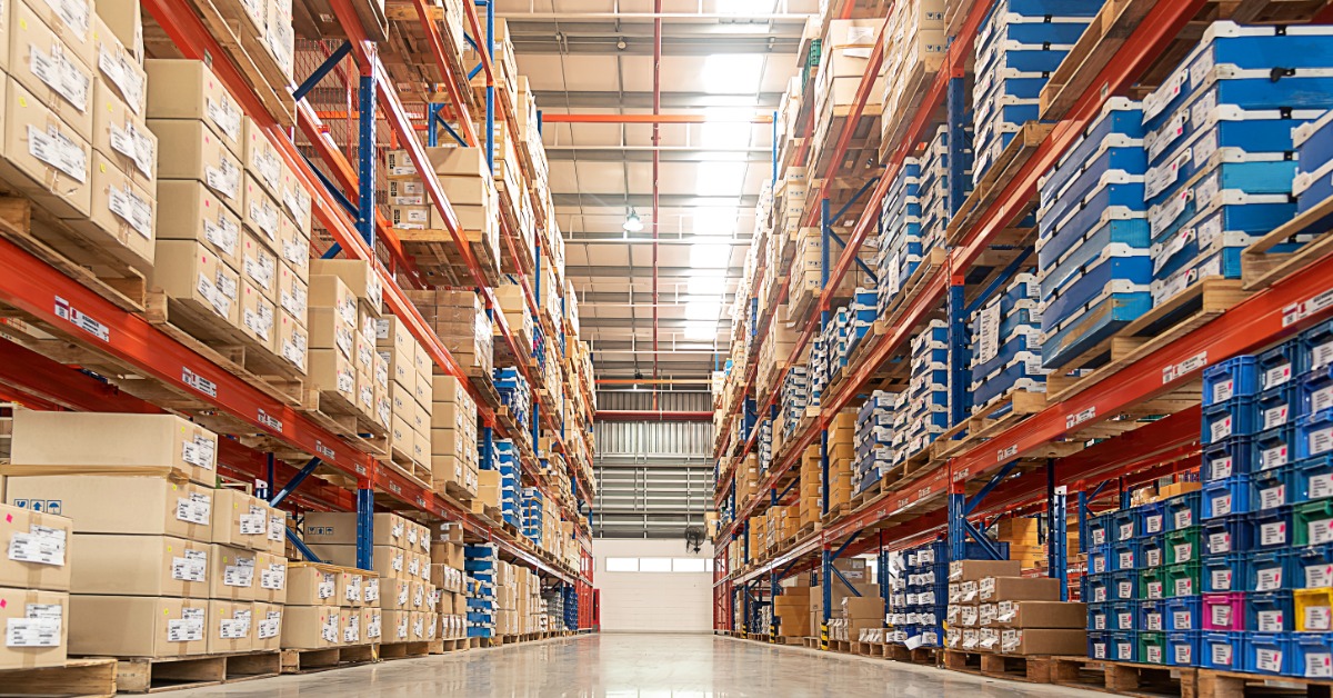 Two rows of floor-to-ceiling shelving in a large industrial space. Boxes, pallets, and blue containers fill the racks.