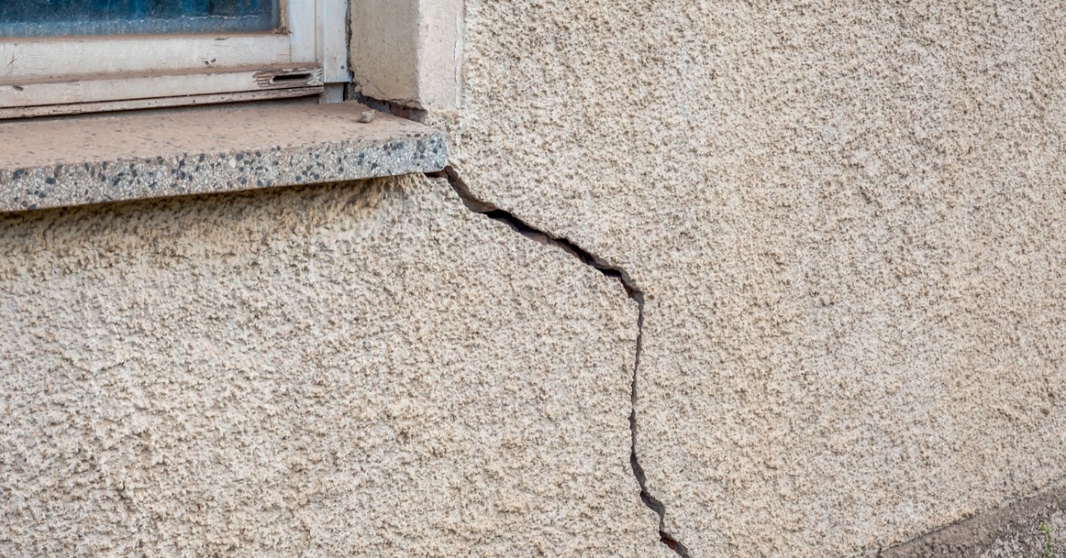 A rough stucco wall has a deep, uneven crack beginning below a window and moving downward at an angle.