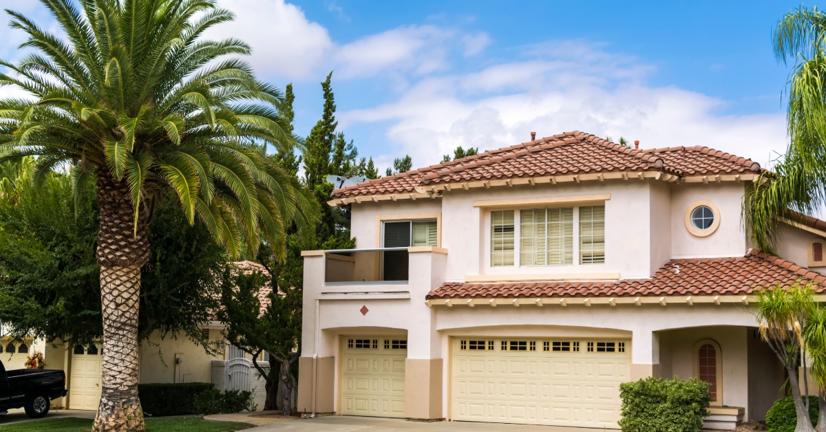 A two-story stucco house has a red tile roof, a wide driveway, and a three-car garage. A palm tree is in the front yard.