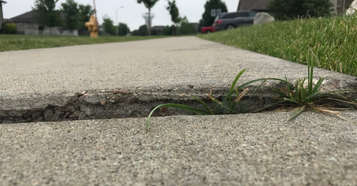 A low-angle view shows two sections of concrete sidewalk with a noticeable height difference. Grass grows in the gap.