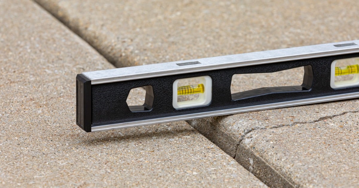 A black-and-silver level rests across two sections of a concrete sidewalk. One slab is higher than the other.