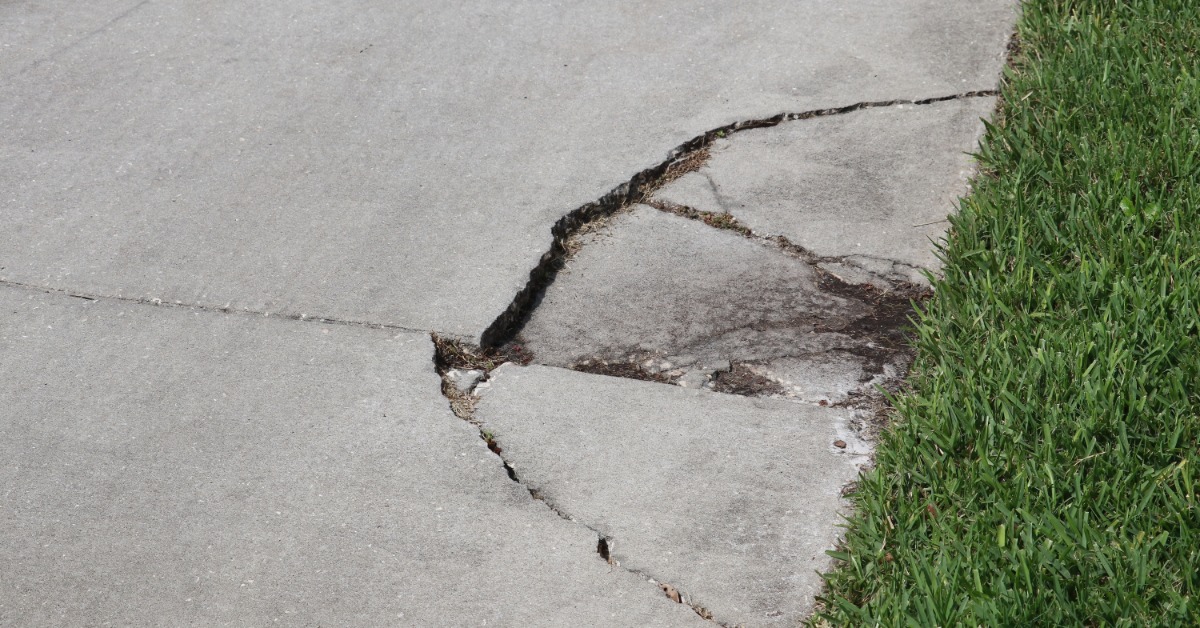 A concrete sidewalk has several wide cracks and broken sections near the edge of a grassy lawn. Some pieces are uneven.