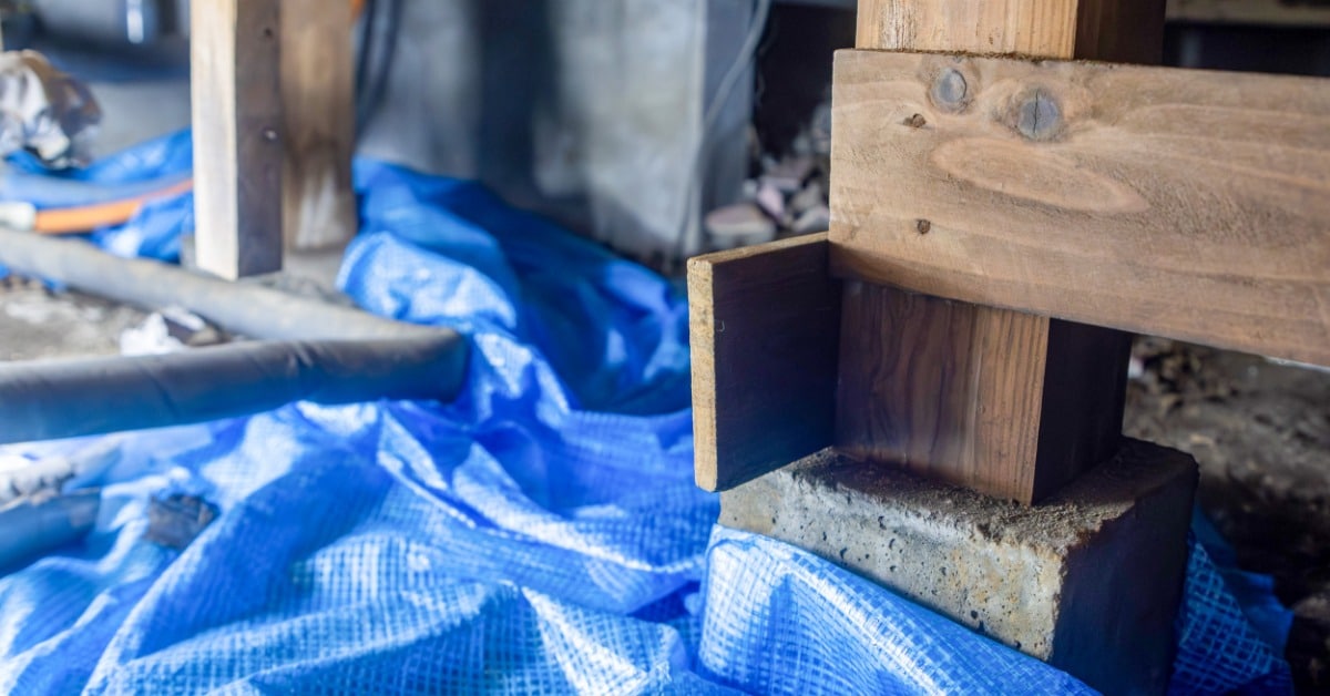 A wooden support post rests on a concrete block in a crawl space. Blue sheeting covers the ground around the post.