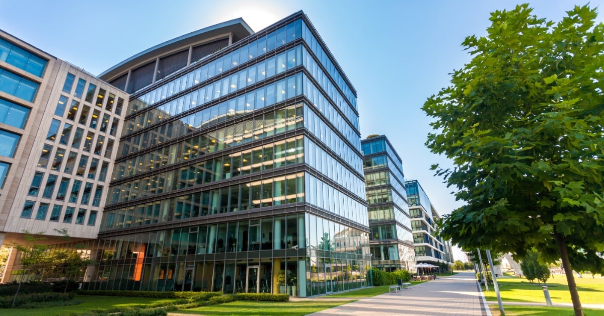 A modern office complex features multi-story buildings with large glass windows, a paved walkway, and landscaping.