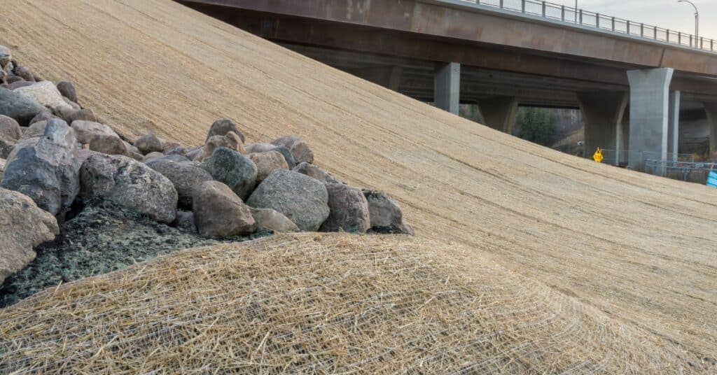 A steep, mat-covered embankment slopes down beside a concrete bridge. Large rocks are clustered in the foreground.