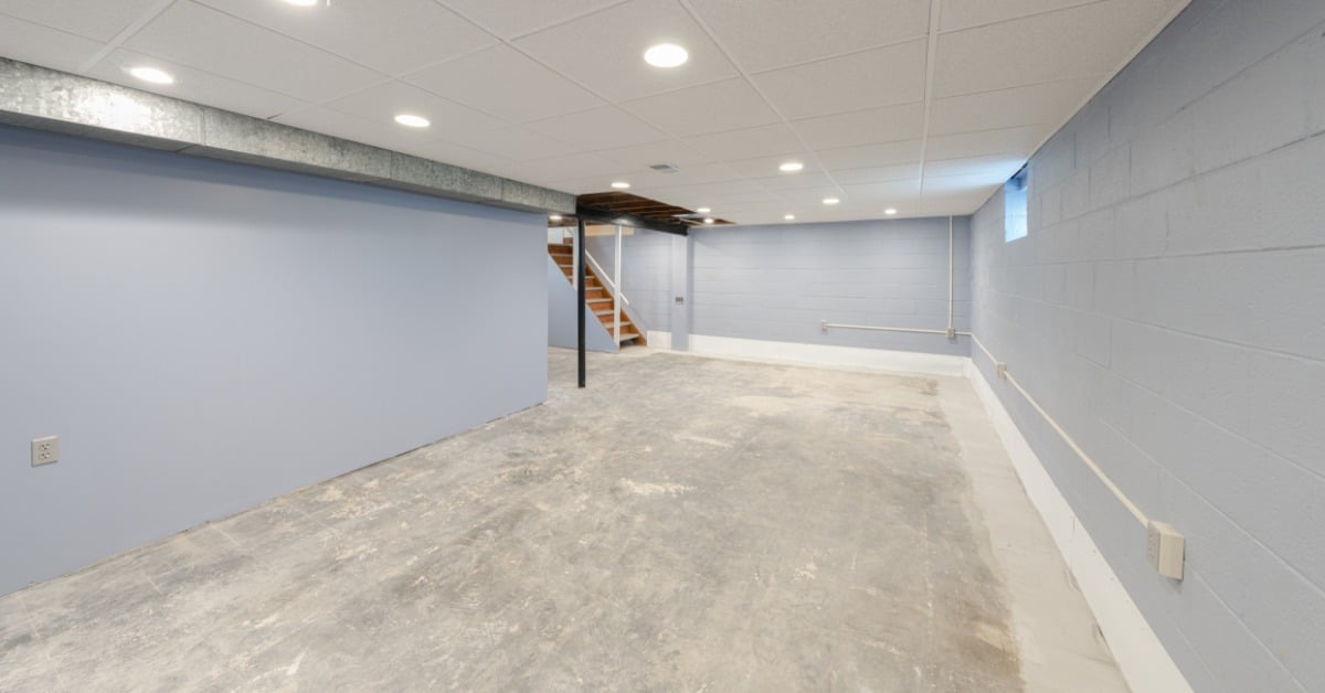 A basement has light blue-gray walls and a bare concrete floor. A staircase and a window are at the far end of the room.