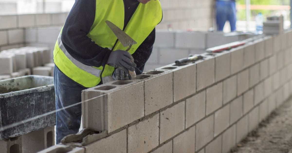 A worker in a reflective vest builds a concrete block wall with a masonry trowel. Fresh mortar is between the blocks.