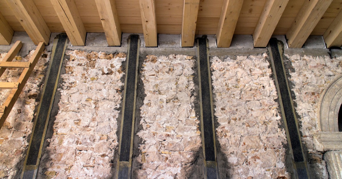 Several vertical black carbon-fiber straps are attached to a rough stone wall beneath a ceiling of exposed wood beams.