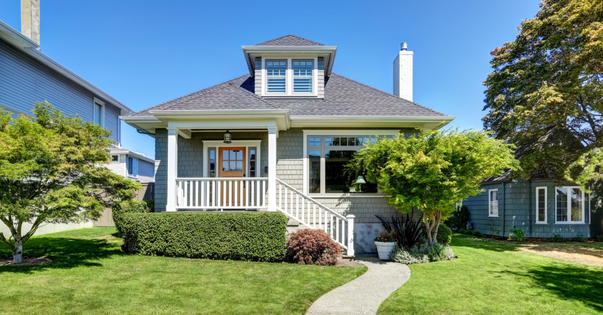 A single-story gray home has a small upper dormer and white porch railings. The front yard is landscaped.
