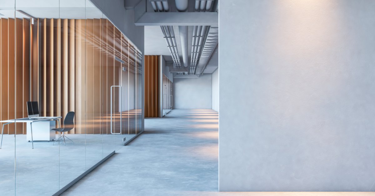 A modern office has concrete floors, glass walls, exposed ceiling pipes, and a workspace behind a glass partition.