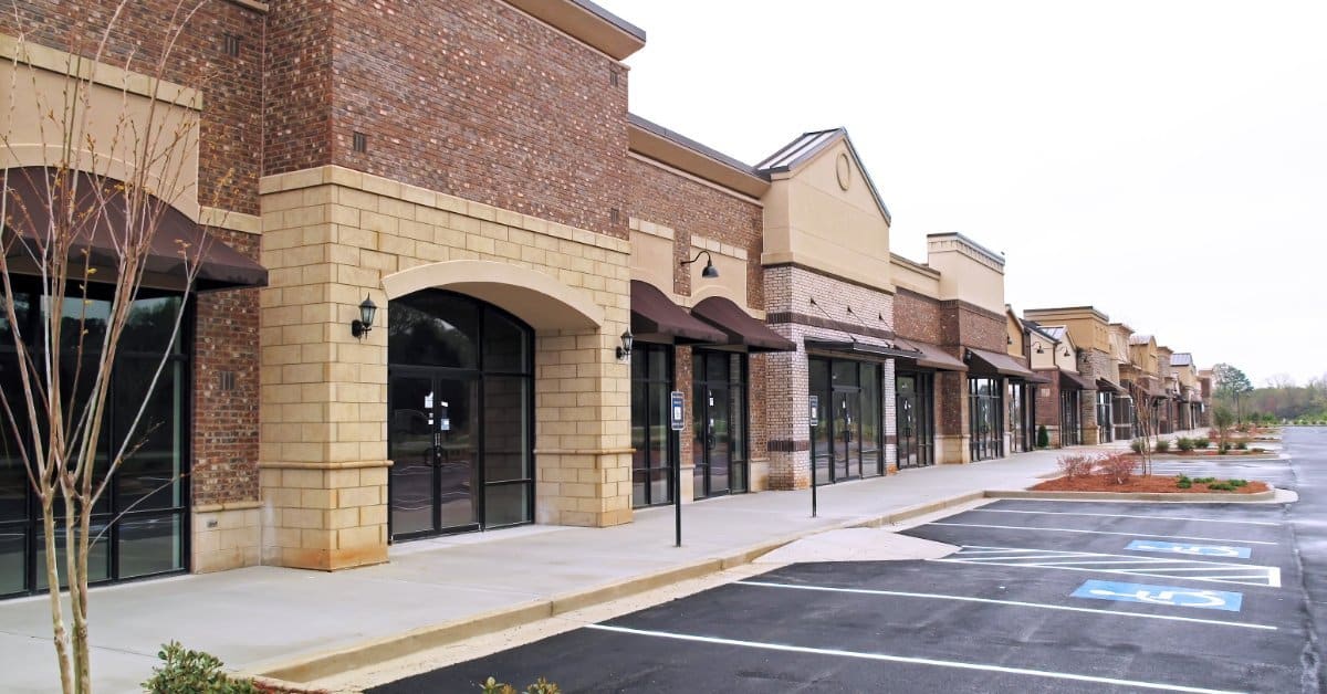 A shopping center features brick and stone storefronts with dark awnings. An empty asphalt parking lot sits in front.