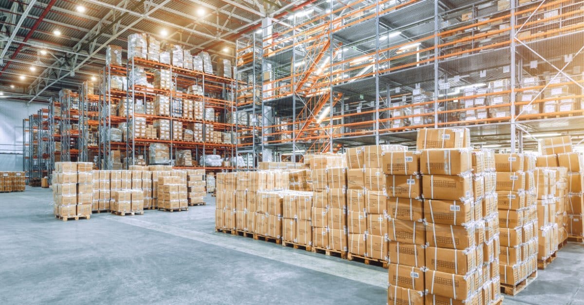 Tall storage racks filled with boxes and wrapped pallets fill a large warehouse. The concrete floor is smooth.